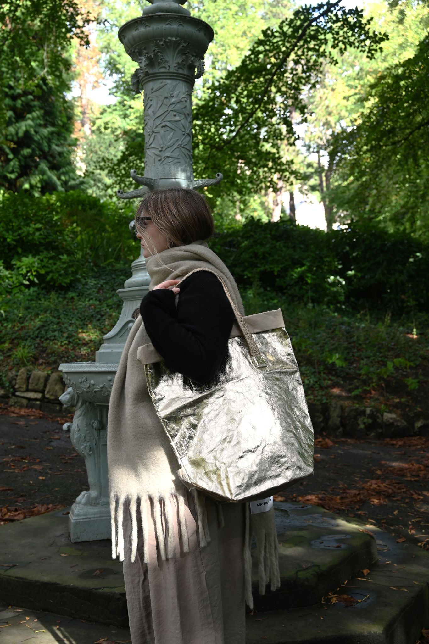 Person holding a large metallic pale gold bag in a park-like setting with trees and a stone structure.