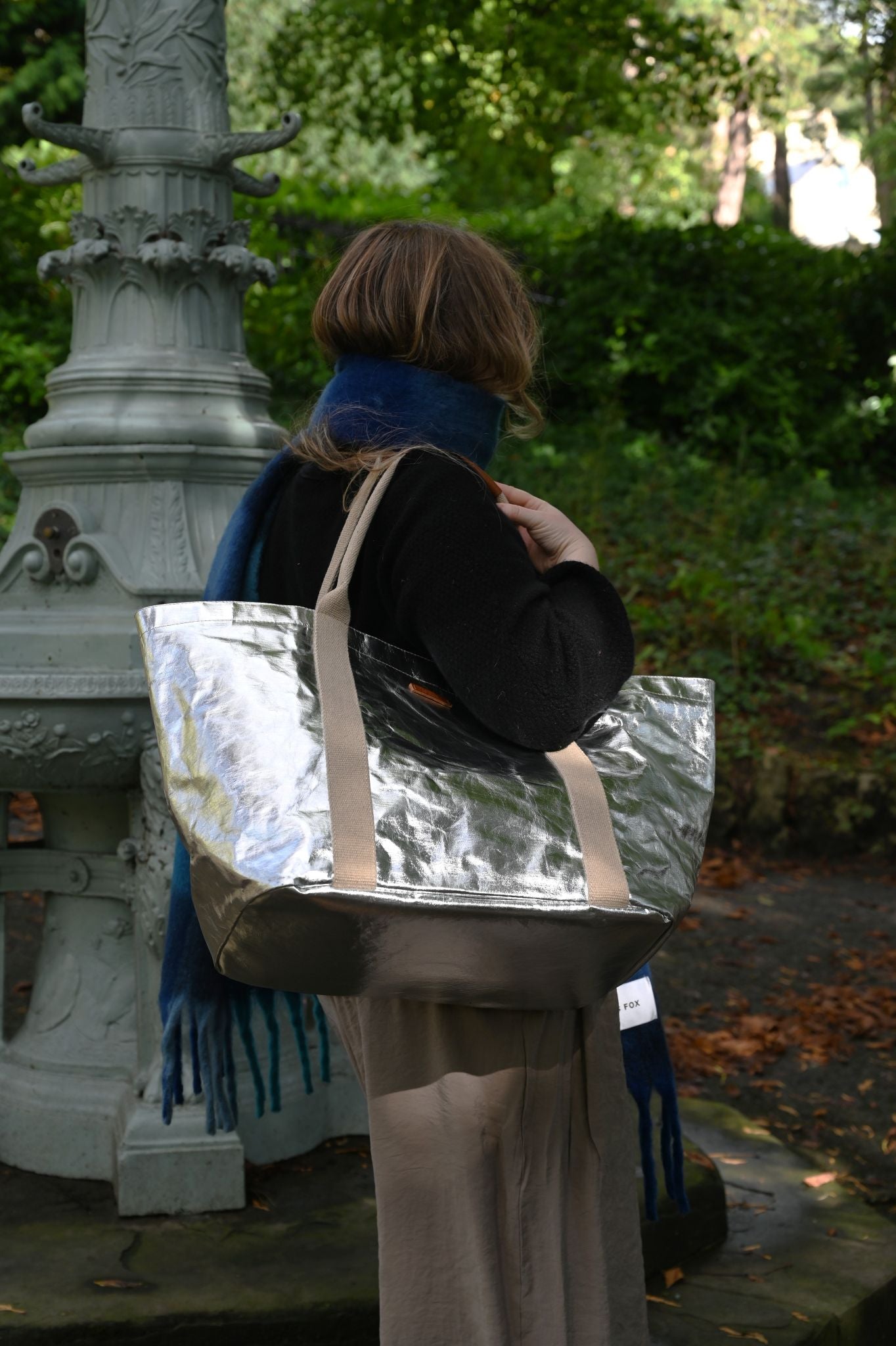 Person holding a metallic large silver tote bag in an outdoor setting with greenery.