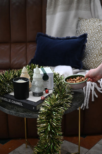 Small round table with decorative items including LED glass christmas trees, give us a snog candle, green and gold tinsel, and a bowl of nuts, with a person's hand reaching in.