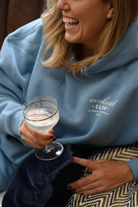 Person wearing a blue sweatshirt with text, holding a glass of champagne, and interacting with another person.