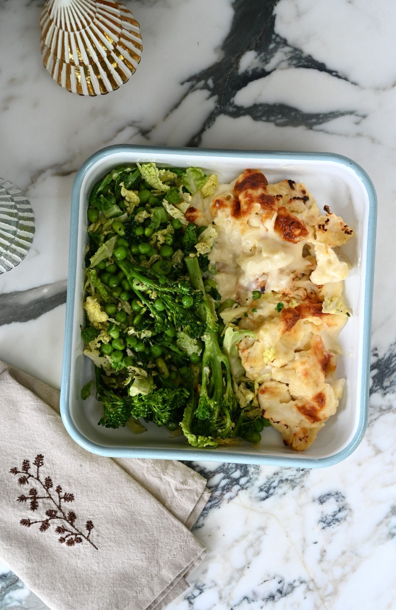 Square enamel tray of food with green vegetables and a cheesy dish on a marble surface