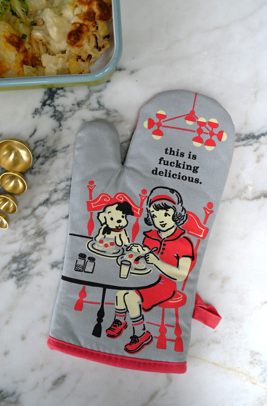Oven mitt with graphic design and 'this is fucking delicious' text on a marble surface with food.