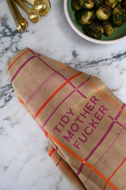 Beige tea towel with plaid pattern and tidy mother fucker text on a marble surface with brussels sprouts and gold measuring spoons.