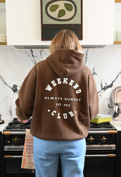 Person wearing a brown hoodie with 'Weekend Always Sunday Club' text in a kitchen.