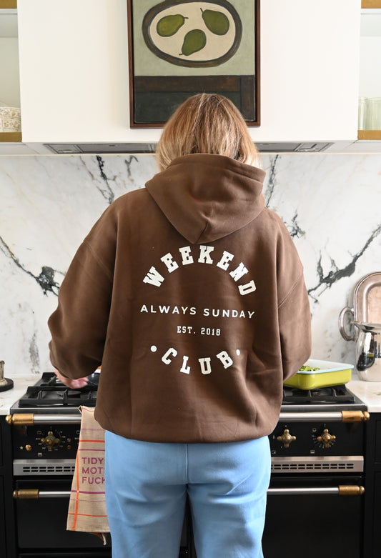 Person wearing a brown hoodie with 'Weekend Always Sunday Club' text in a kitchen.