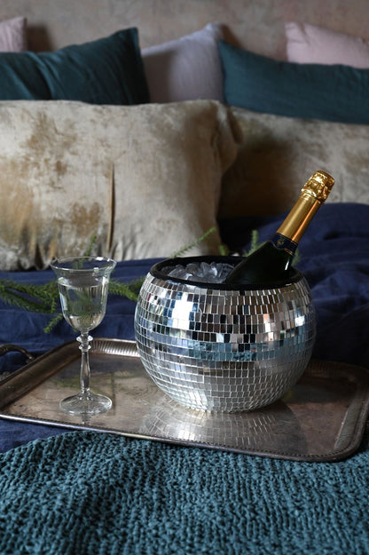 Champagne bottle in a disco ball on a tray with a glass, set against a textured surface with pillows.