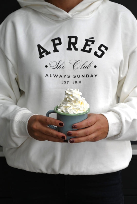 Person wearing a white hoodie with 'APRèS Ski Club' text, holding a hot chocolate enamel mug with whipped cream.