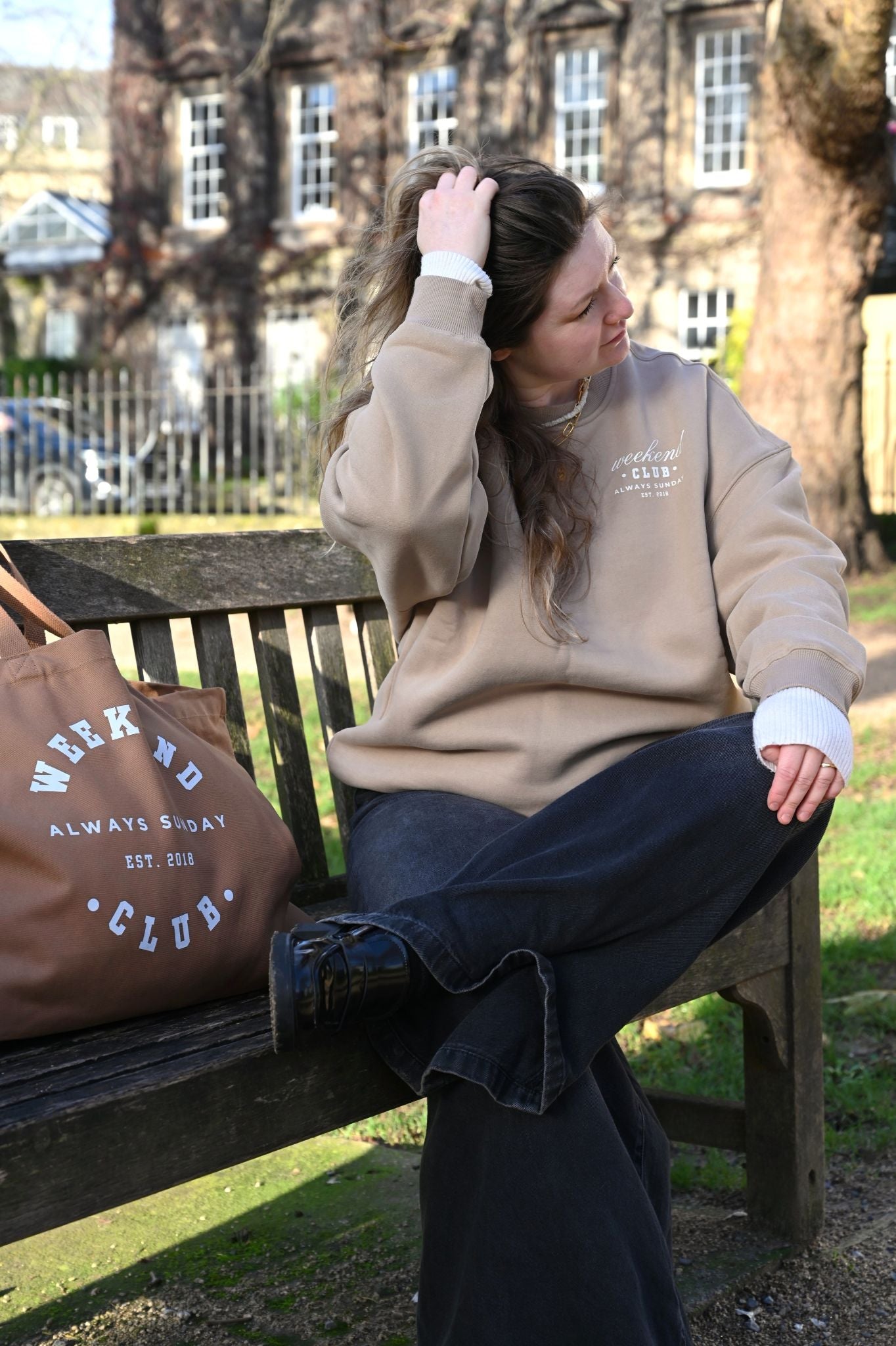 Woman sitting on a park bench wearing a beige sweatshirt and dark jeans, with a brown bag next to her.