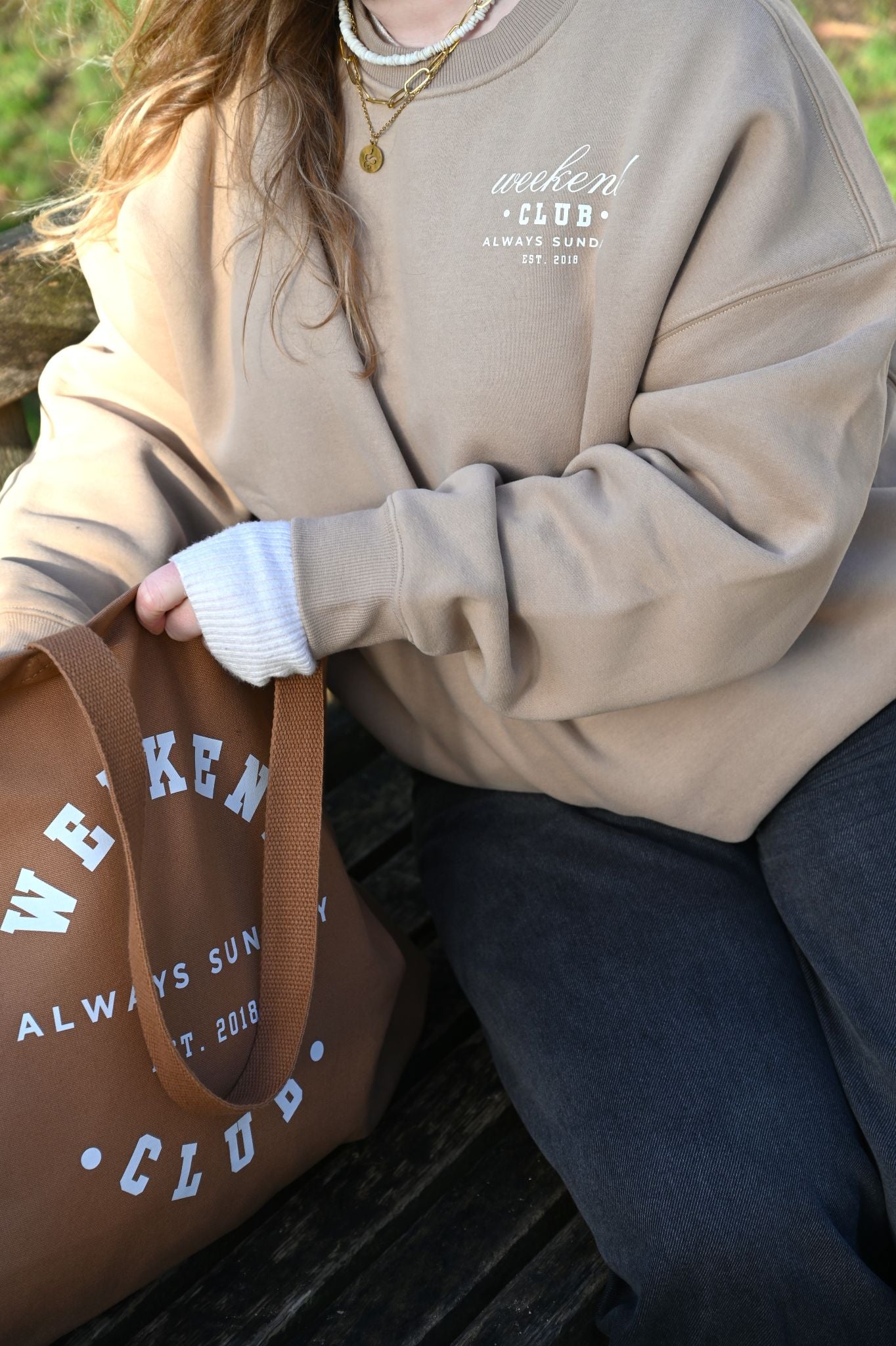 Person wearing a beige sweatshirt with 'Weekend Club' text, holding a brown tote bag with matching branding.