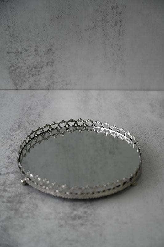 Ornate silver mirrored round tray with filigree edging holding a candle and mug on a dark table.