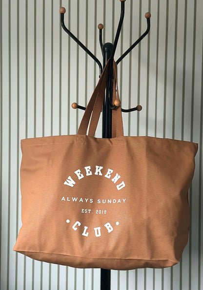 Brown tote bag with 'Weekend Always Sunday Club' text hanging on a coat rack against a striped wall.