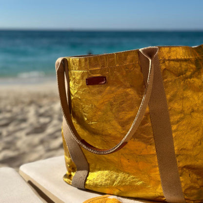 A large lemon gold tote bag with leather details, placed on a sandy beach with the ocean in the background.