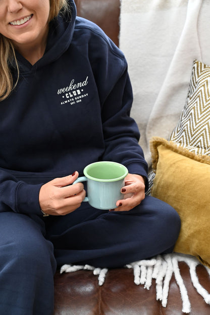 Person wearing a blue weekend club hoodie, holding an enamel mug, sitting on a couch.